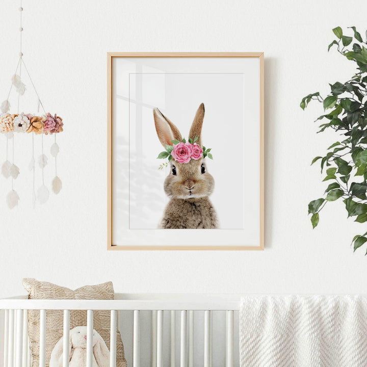 A framed picture of a bunny wearing a flower crown is hanging on a white wall above a white crib.