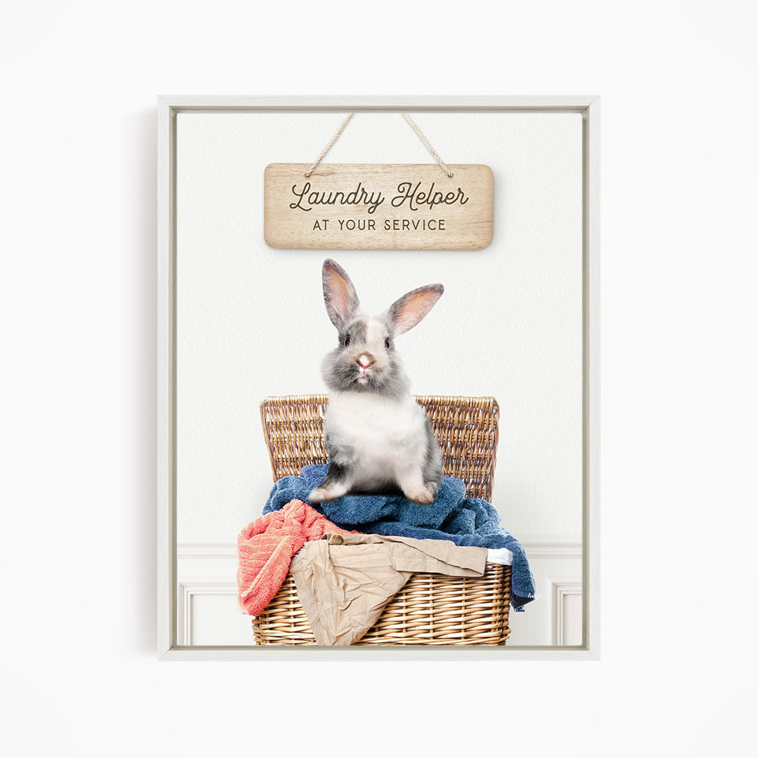 A gray and white rabbit sitting in a wicker laundry basket filled with clothes, with a wooden sign above that reads "Laundry Helper at your service".