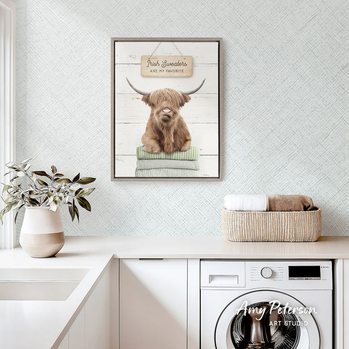 a framed picture of a Highland cow, a breed of cattle known for their long, shaggy hair, sitting on a stack of towels. The towel stack is placed on a white countertop, and there is a basket of towels nearby. The wall behind the countertop has a textured pattern, and there is a potted plant on the countertop.