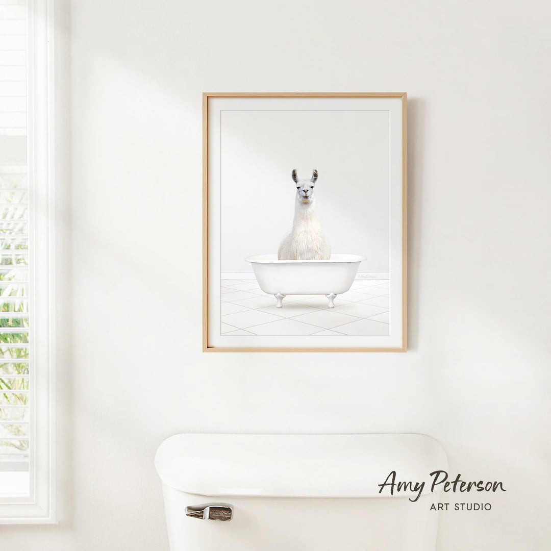A framed image of a white llama sitting in a white bathtub is hanging on a white wall above a white toilet.
