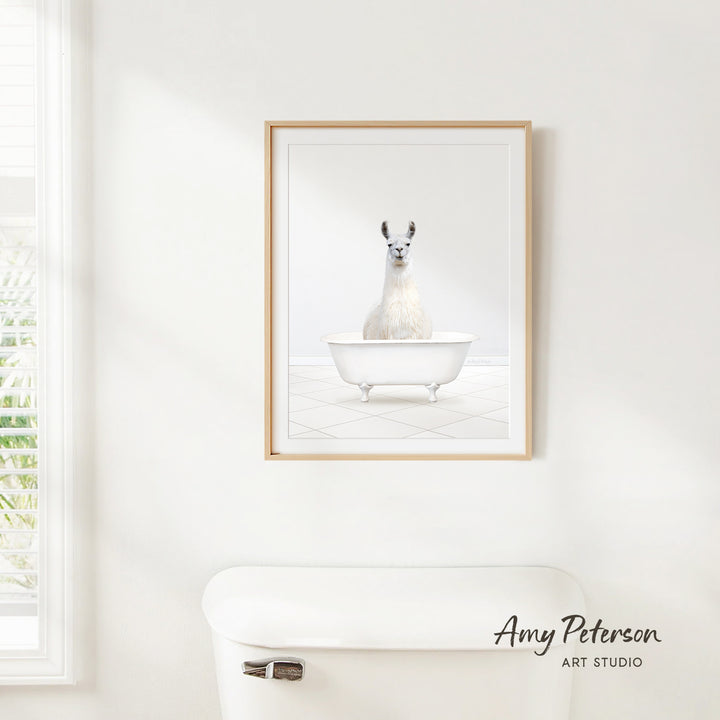 A framed image of a white llama sitting in a white bathtub is hanging on a white wall above a white toilet.