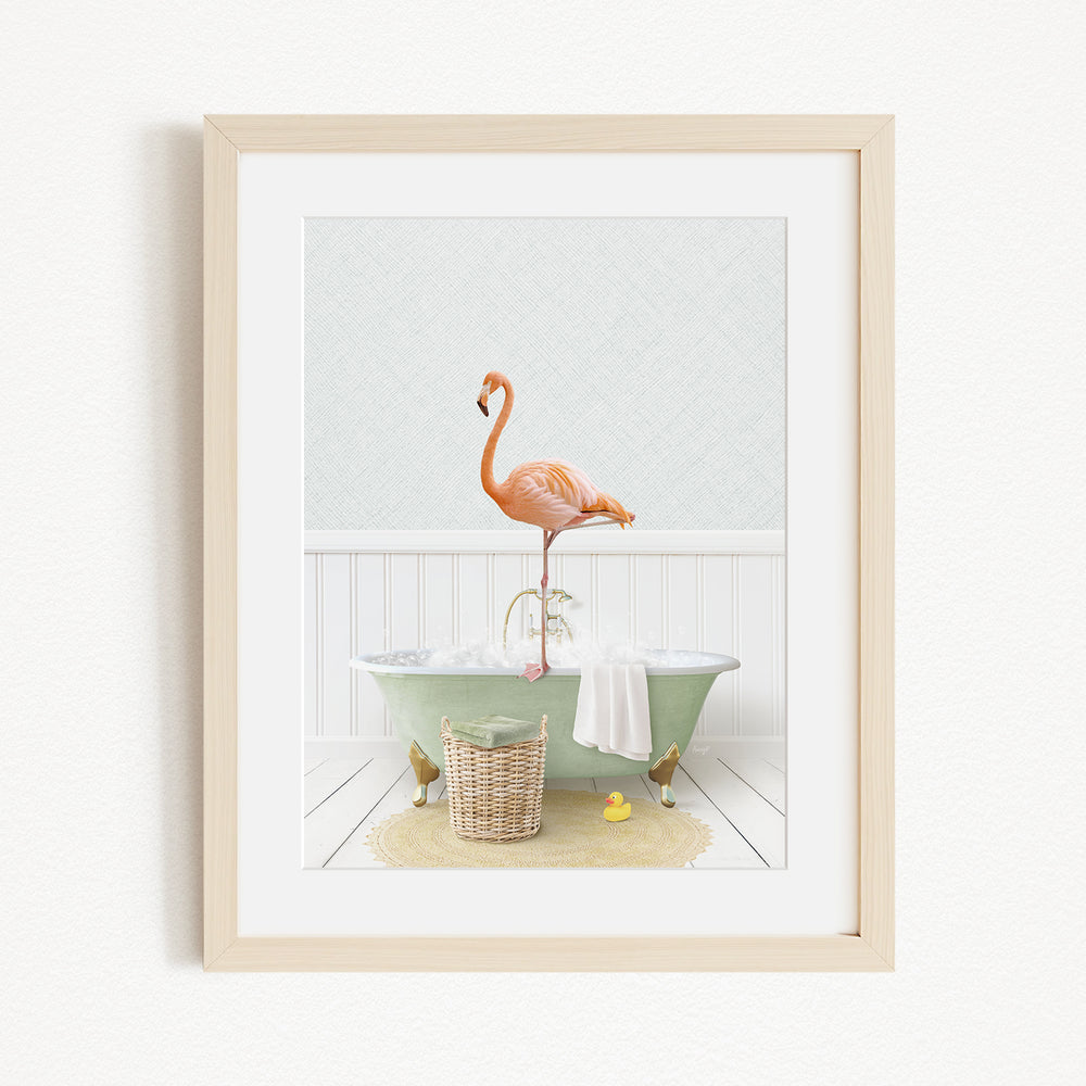 A framed image depicting a pink flamingo standing on the edge of a green bathtub, with a basket of bath toys and a yellow rubber duck nearby.