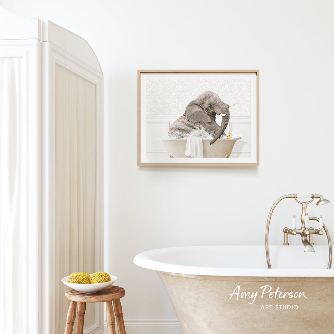 A framed image of an elephant taking a bath in a tub, hanging on a white wall above a wooden stool with a bowl of fruit on it.