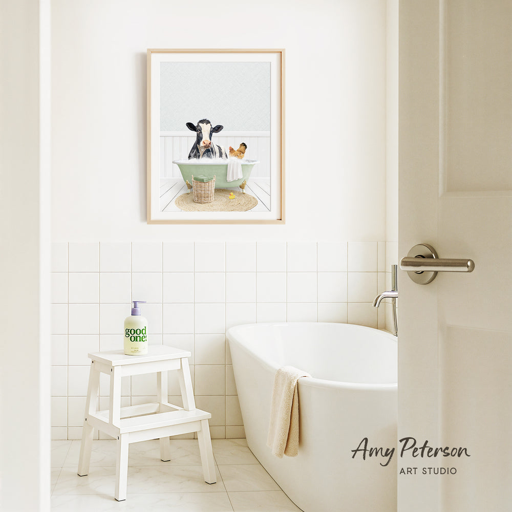 A framed image of a cow sitting in a bathtub is displayed on the wall above a white bathtub in a bathroom.