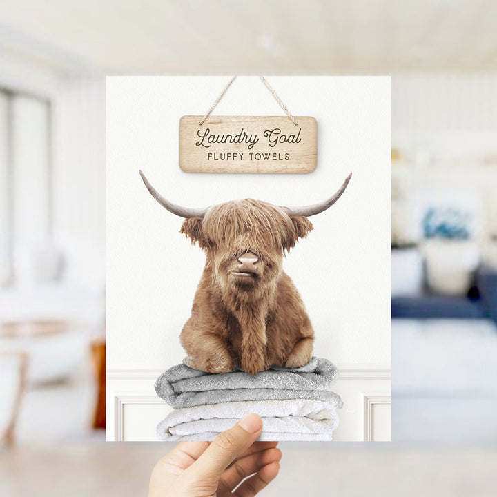 A hand holding a framed print featuring a brown cow with long horns sitting on a stack of fluffy towels, with a sign above that says "Laundry Goal Fluffy Towels".
