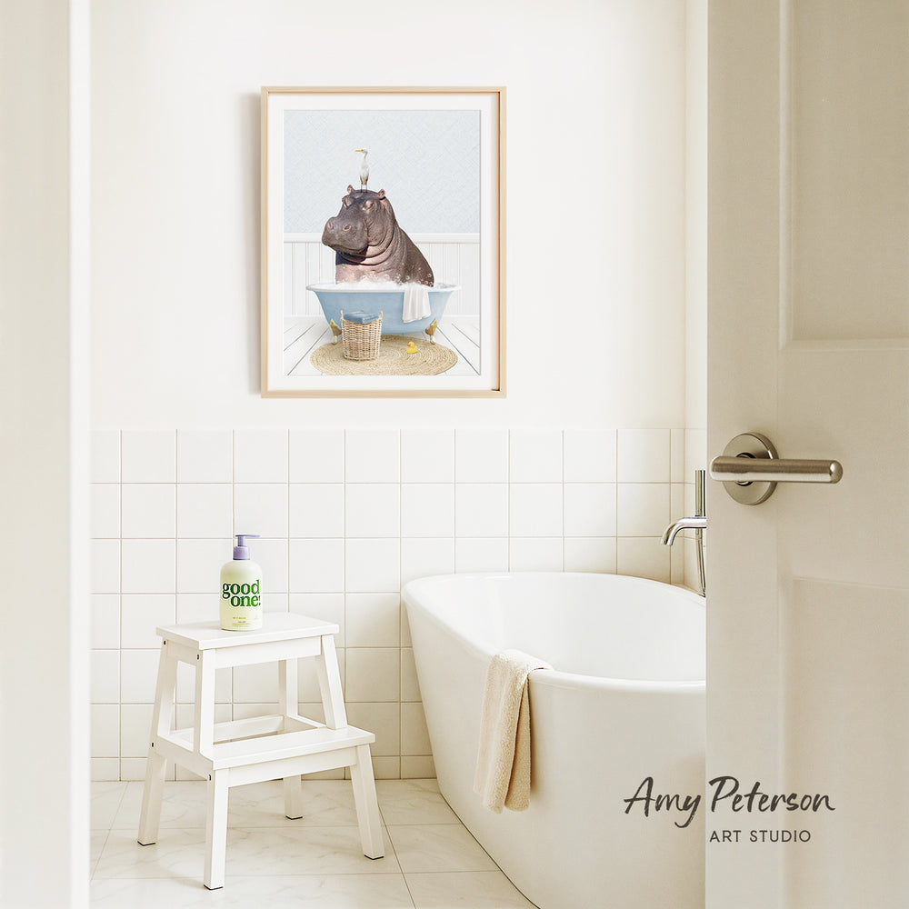 A framed image of a hippopotamus sitting in a bathtub is displayed on the wall above a white bathtub in a bathroom.