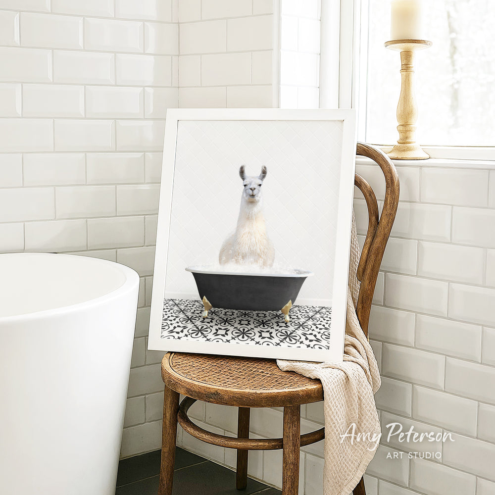 A framed image of a white llama sitting in a black bathtub, surrounded by a patterned tile design, is displayed on a wooden chair in a bathroom setting.