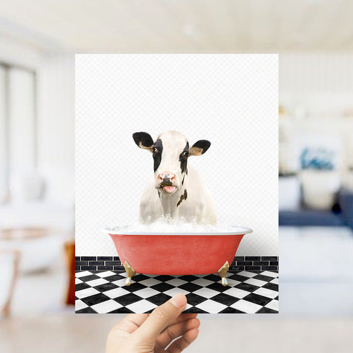 A hand holds up a framed image of a cow sitting in a red bathtub filled with water, creating a whimsical and humorous scene.