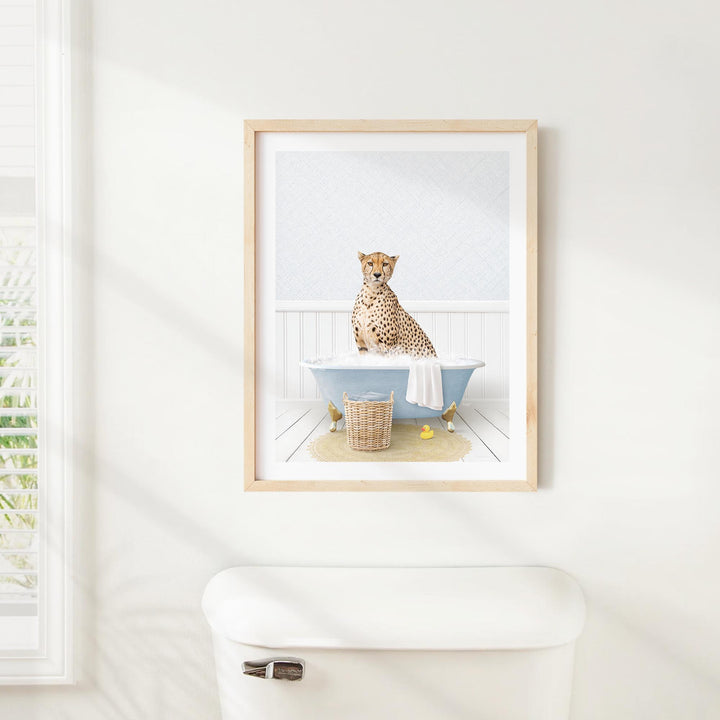 A cheetah sitting in a bathtub, with a basket of bath toys nearby, is the central focus of the framed image displayed on the wall.