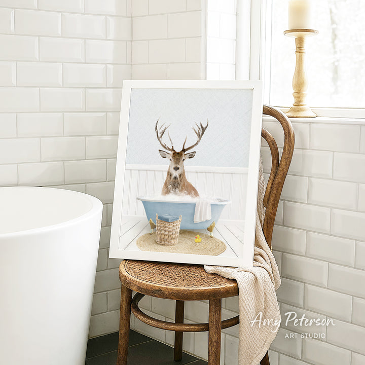 A framed image of a deer sitting in a bathtub, with a towel and basket nearby, is displayed on a wooden chair in a bathroom setting.