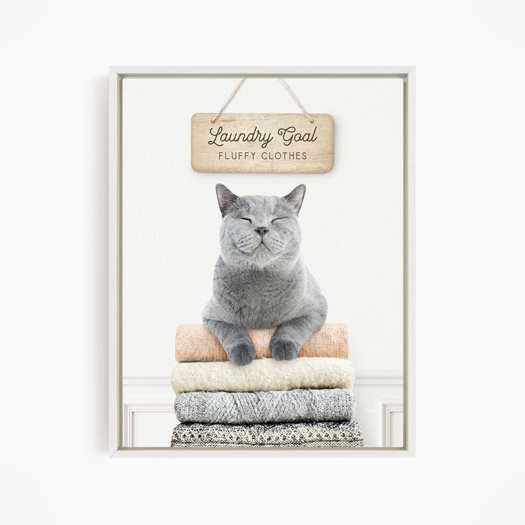 A gray cat sitting on a stack of folded clothes, with a wooden sign above that says "Laundry Goal Fluffy Clothes".