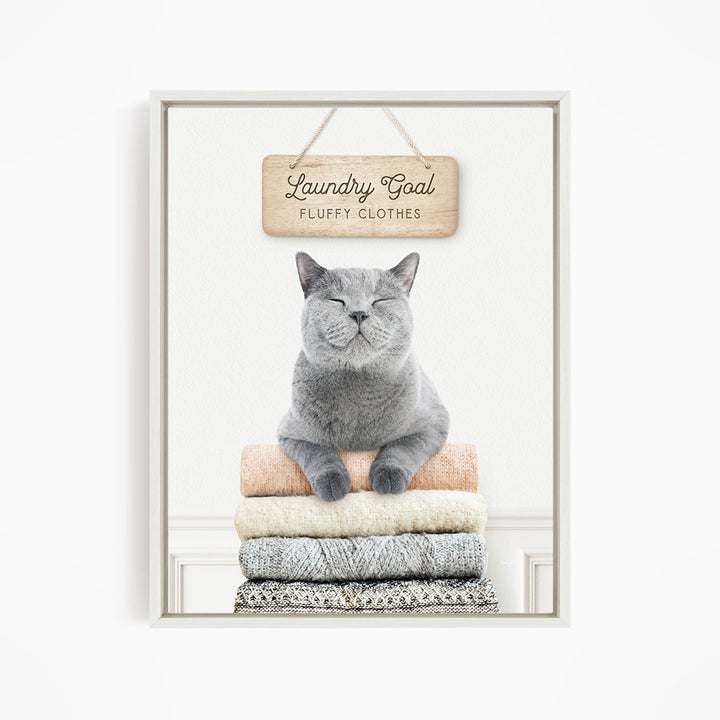 A gray cat sitting on a stack of folded clothes, with a wooden sign above that says "Laundry Goal Fluffy Clothes".