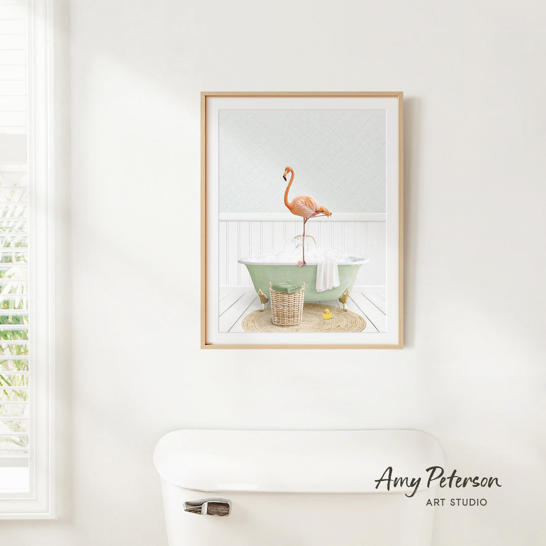 A framed image of a pink flamingo standing in a green bathtub, with a basket of bath toys nearby, is displayed on a white wall in a bathroom.