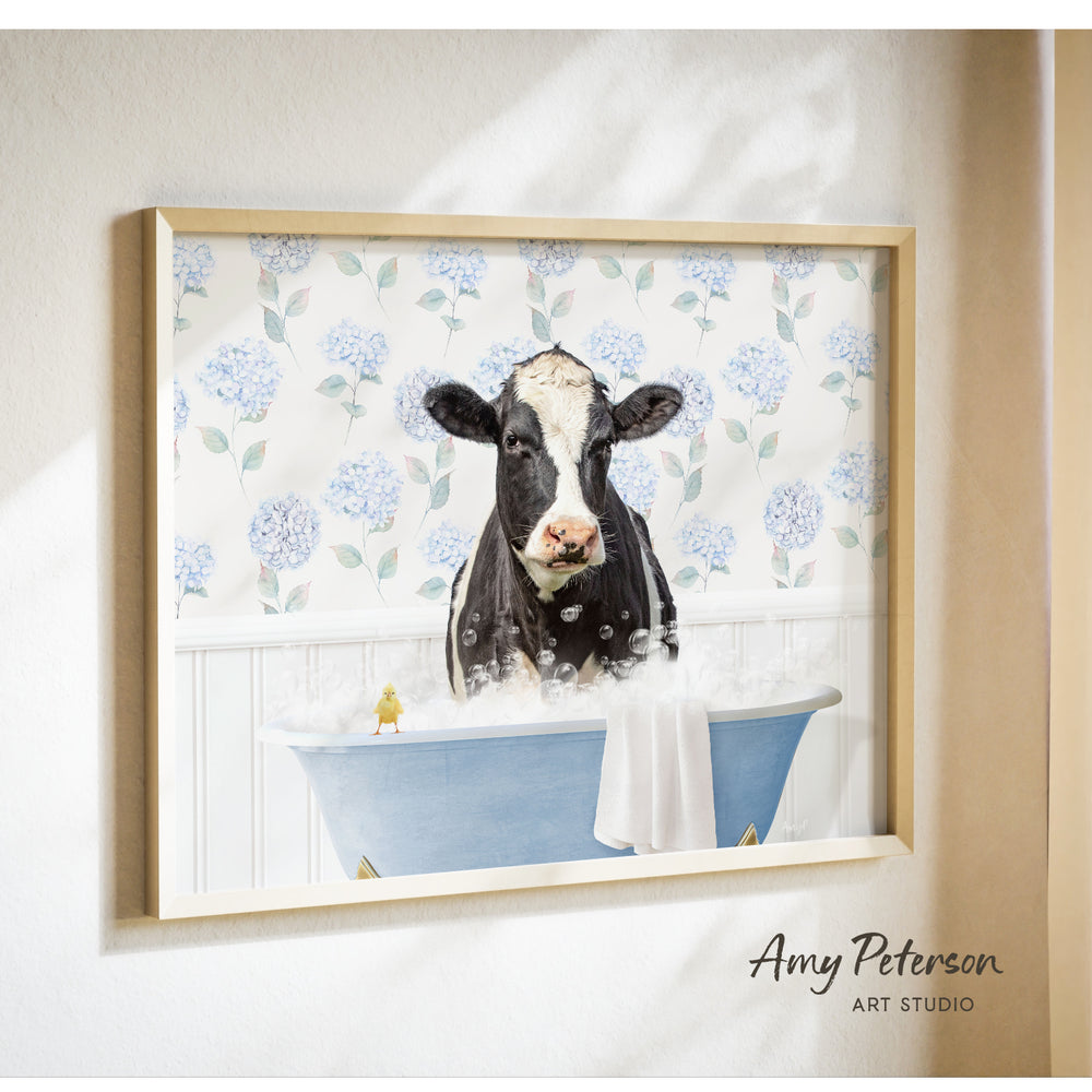 A framed image of a black and white cow sitting in a blue bathtub filled with bubbles, surrounded by a floral pattern on the wall behind it.