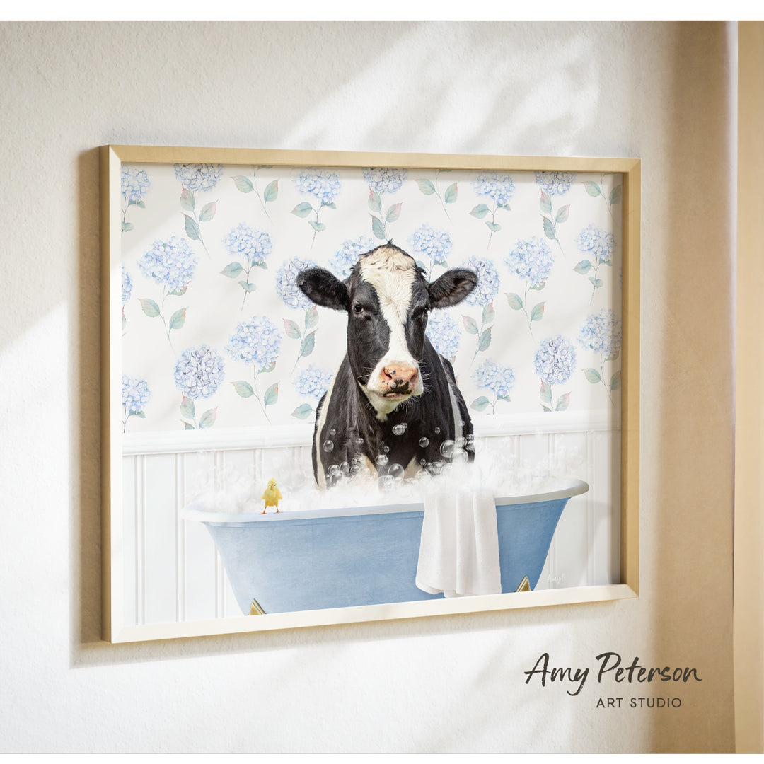 A framed image of a black and white cow sitting in a blue bathtub filled with bubbles, surrounded by a floral pattern on the wall behind it.