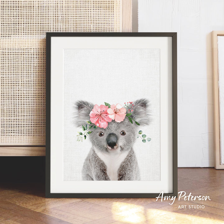 A framed print of a koala bear wearing a floral headpiece is displayed on a wooden floor against a white wall.