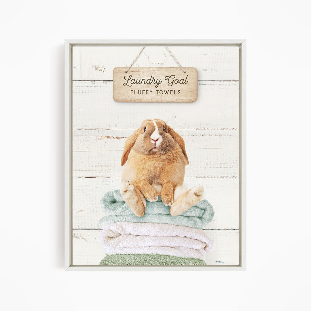A wooden sign with the text "Laundry Goal Fluffy Towels" hangs above a stack of folded towels, with a rabbit sitting on top of the towels.