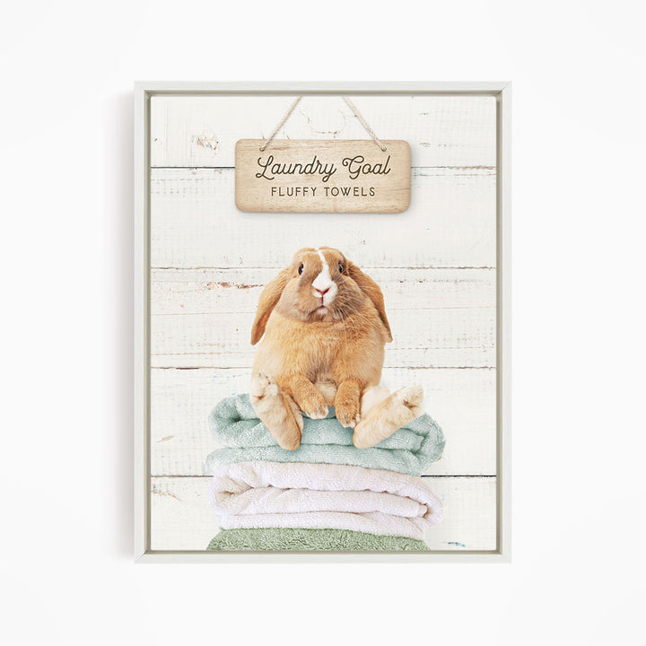 A wooden sign with the text "Laundry Goal Fluffy Towels" hangs above a stack of folded towels, with a rabbit sitting on top of the towels.