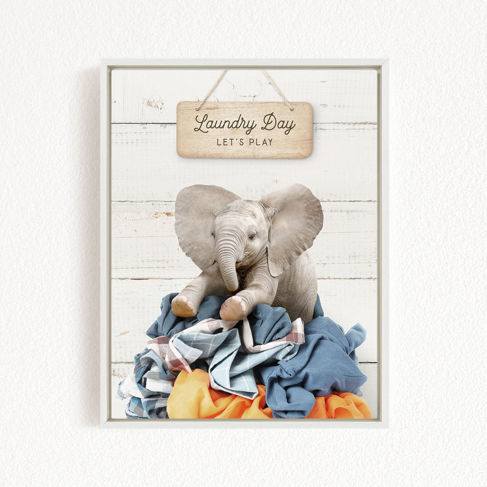 A framed image of a stuffed elephant sitting atop a pile of laundry, with a wooden sign above that says "Laundry Day Let's Play".