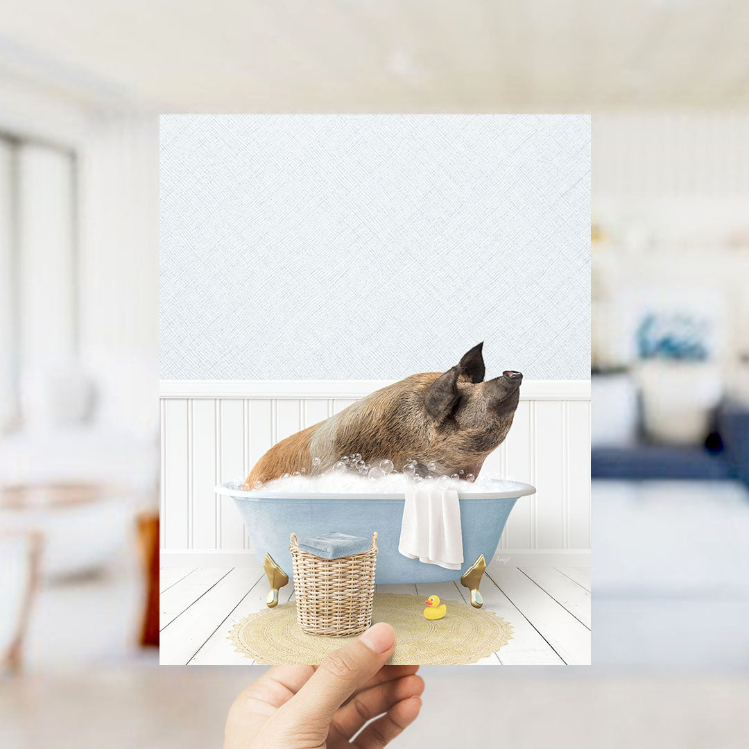 A hand holds up a framed image depicting a pig sitting in a bathtub filled with bubbles, surrounded by a yellow rubber duck and a white towel.