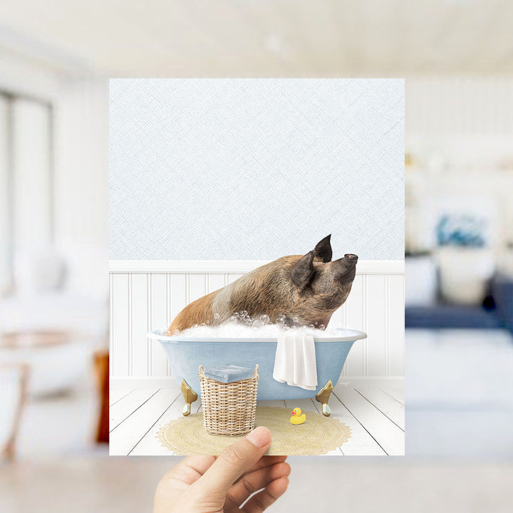 A hand holds up a framed image depicting a pig sitting in a bathtub filled with bubbles, surrounded by a yellow rubber duck and a white towel.