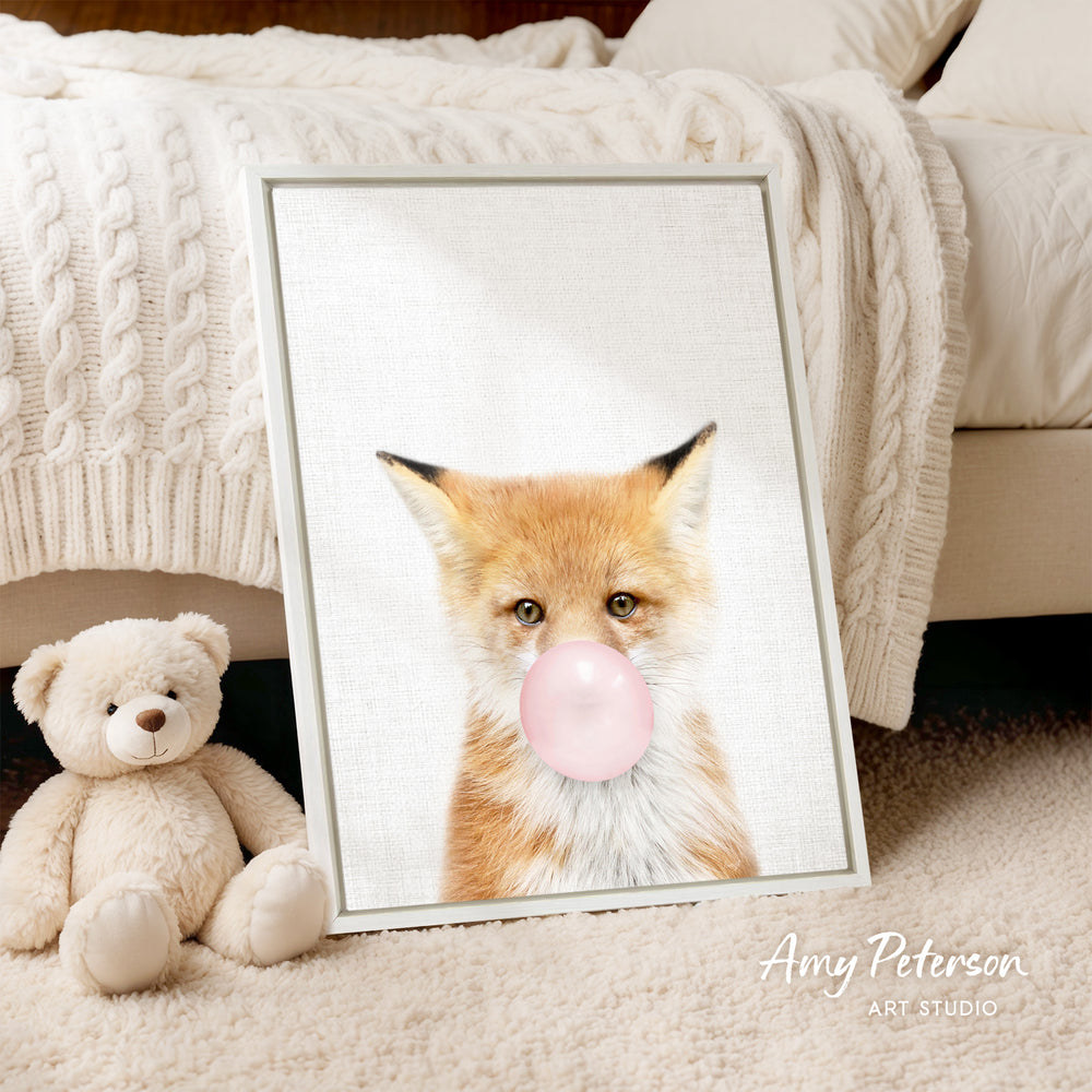 A framed print of a fox blowing a bubble with a pink bubble gum bubble is displayed on a bed, accompanied by a teddy bear.