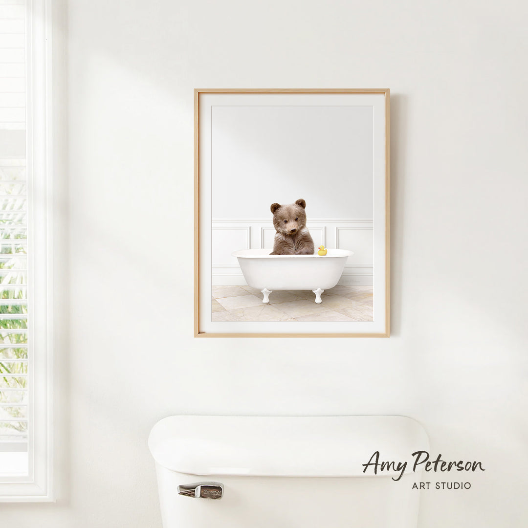 A framed image of a brown bear sitting in a white bathtub, with a yellow rubber duck floating in the water.