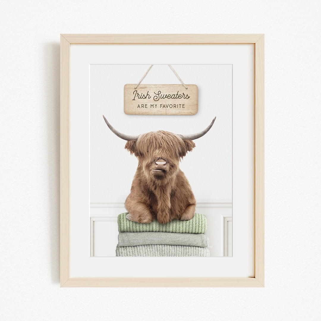 A framed image of a brown cow with long horns, sitting on a stack of green and gray sweaters, with a wooden sign above that says "Irish Sweaters are my favorite".