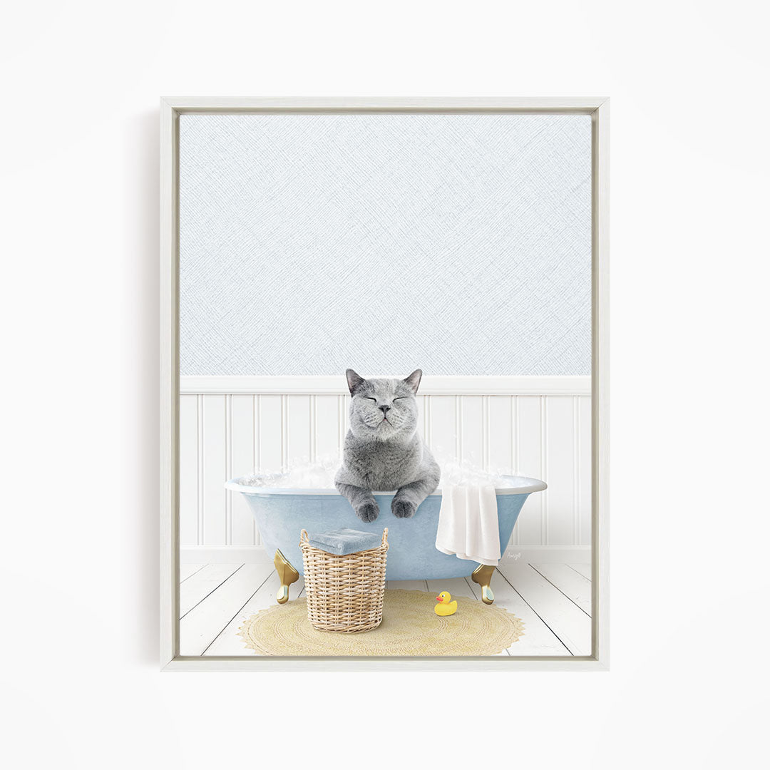 A gray cat sitting in a blue bathtub filled with bubbles, with a yellow rubber duck nearby and a basket of bath toys on the floor.
