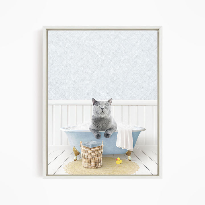 A gray cat sitting in a blue bathtub filled with bubbles, with a yellow rubber duck nearby and a basket of bath toys on the floor.