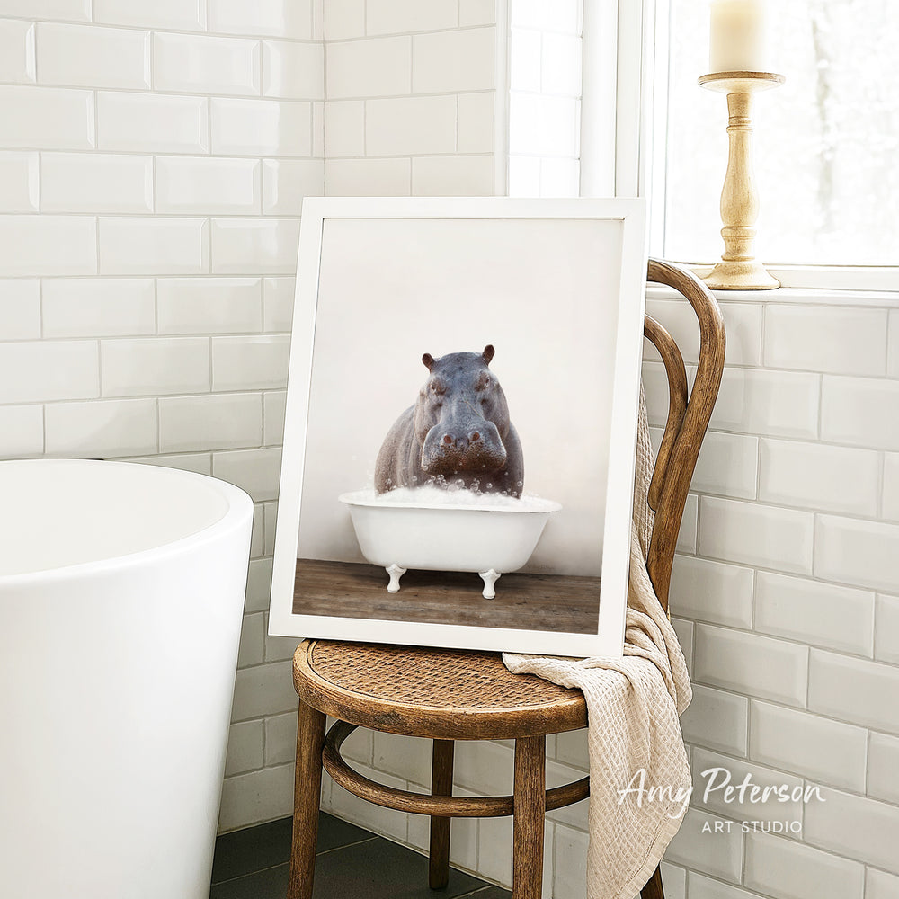 A framed photograph of a hippopotamus sitting in a bathtub is displayed on a wooden chair in a bathroom setting.