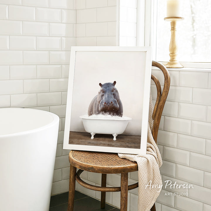 A framed photograph of a hippopotamus sitting in a bathtub is displayed on a wooden chair in a bathroom setting.