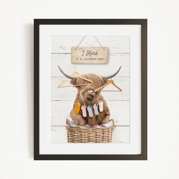 A framed image depicting a Highland cow sitting in a wicker basket, surrounded by clothes and a sign that reads "It's laundry day".