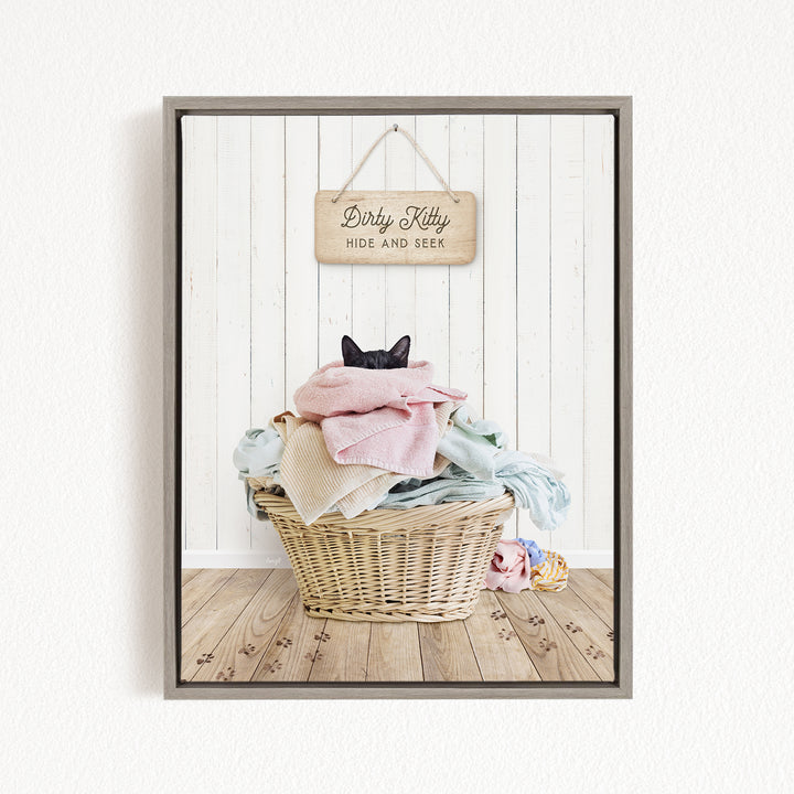 A framed image of a cat sitting in a basket filled with clothes, with a wooden sign above it that says "Dirty Kitty HIDE AND SEEK".
