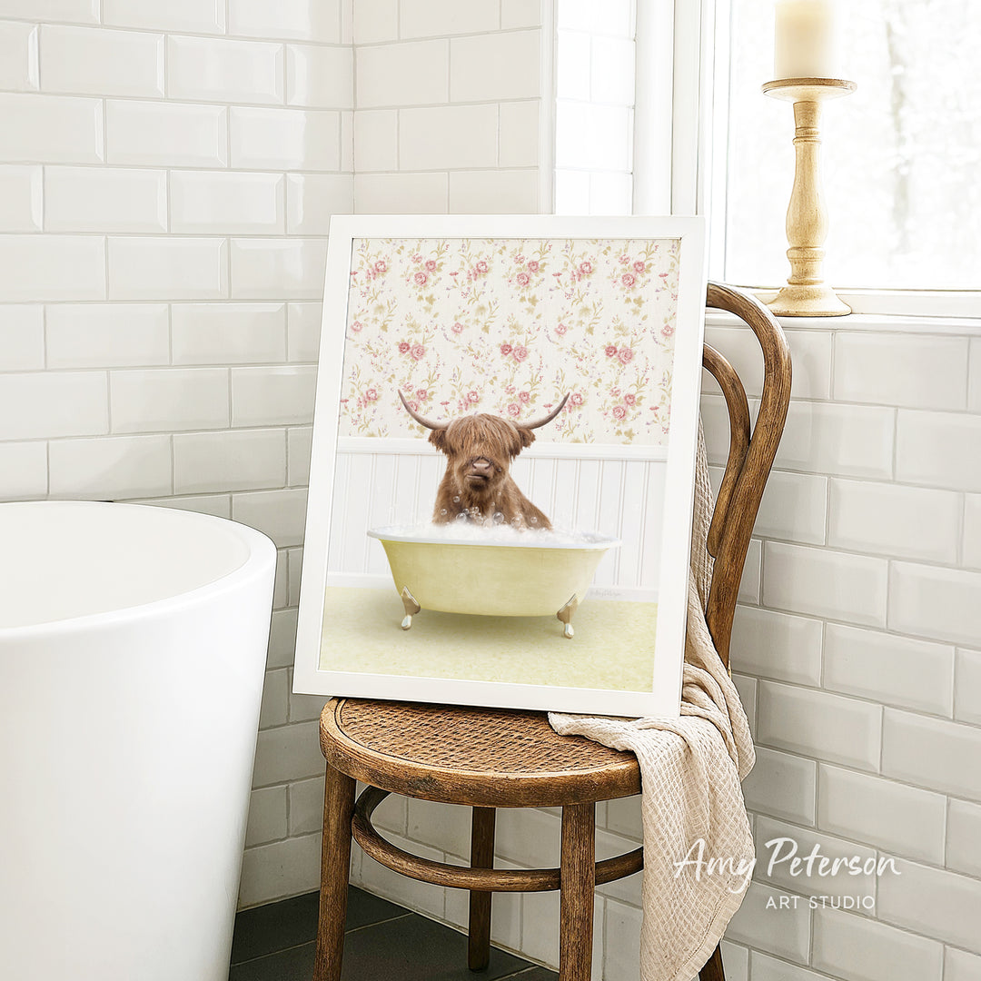 A framed image of a bull with horns sitting in a bathtub, surrounded by a floral pattern, is displayed on a wooden chair in a bathroom setting.