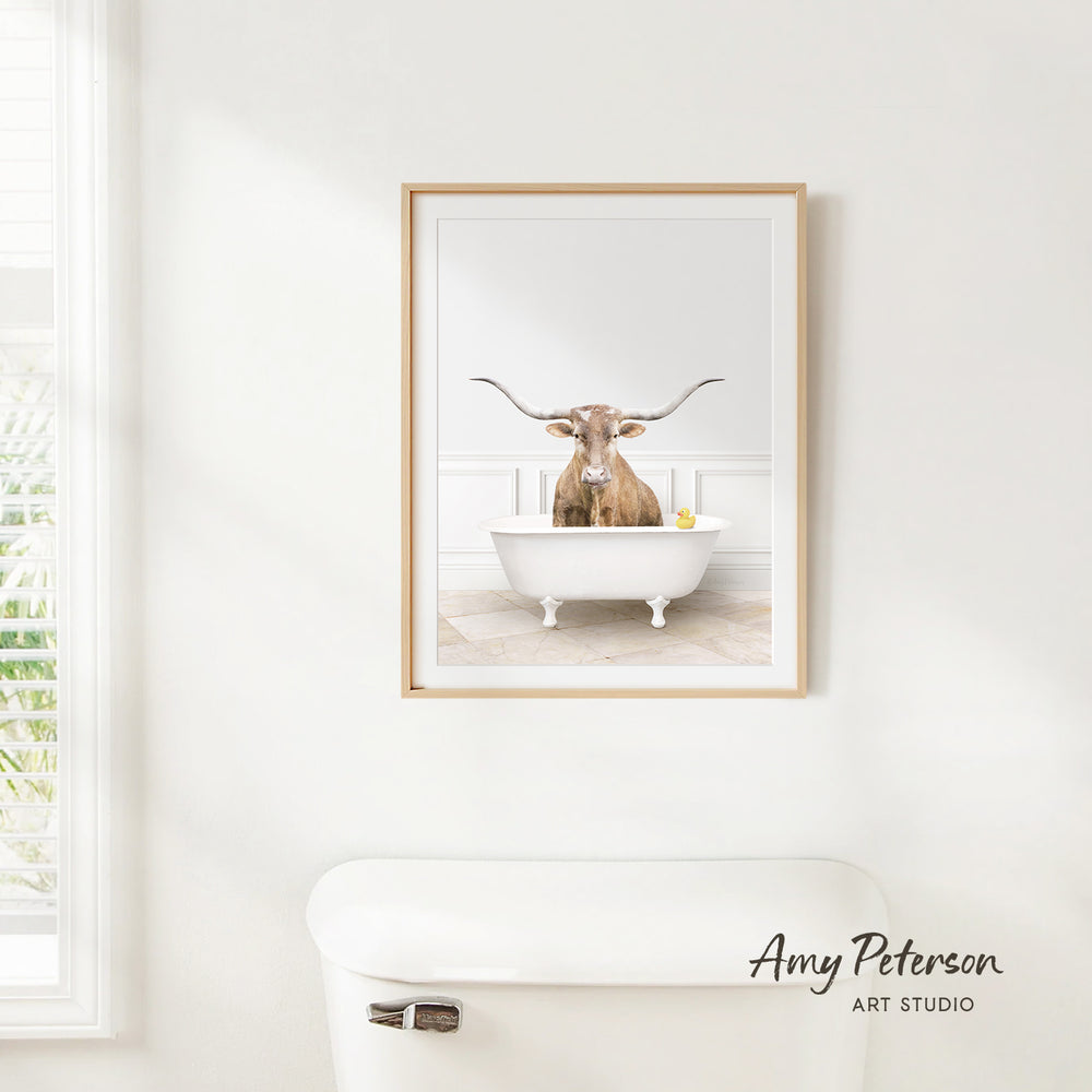 A framed image of a bull with long horns sitting in a white bathtub, with a window and toilet visible in the background.