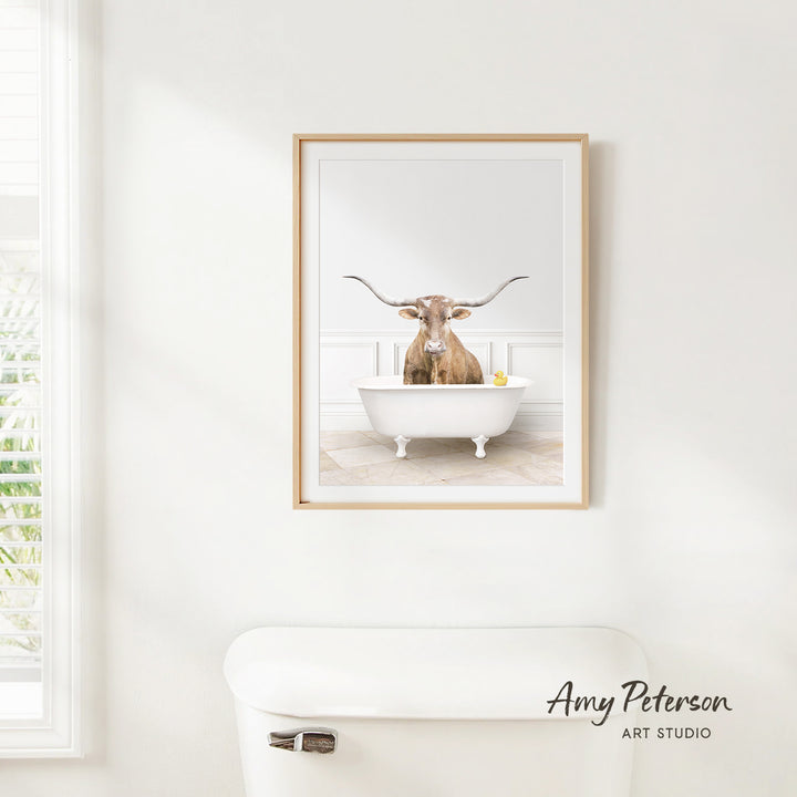 A framed image of a bull with long horns sitting in a white bathtub, with a window and toilet visible in the background.