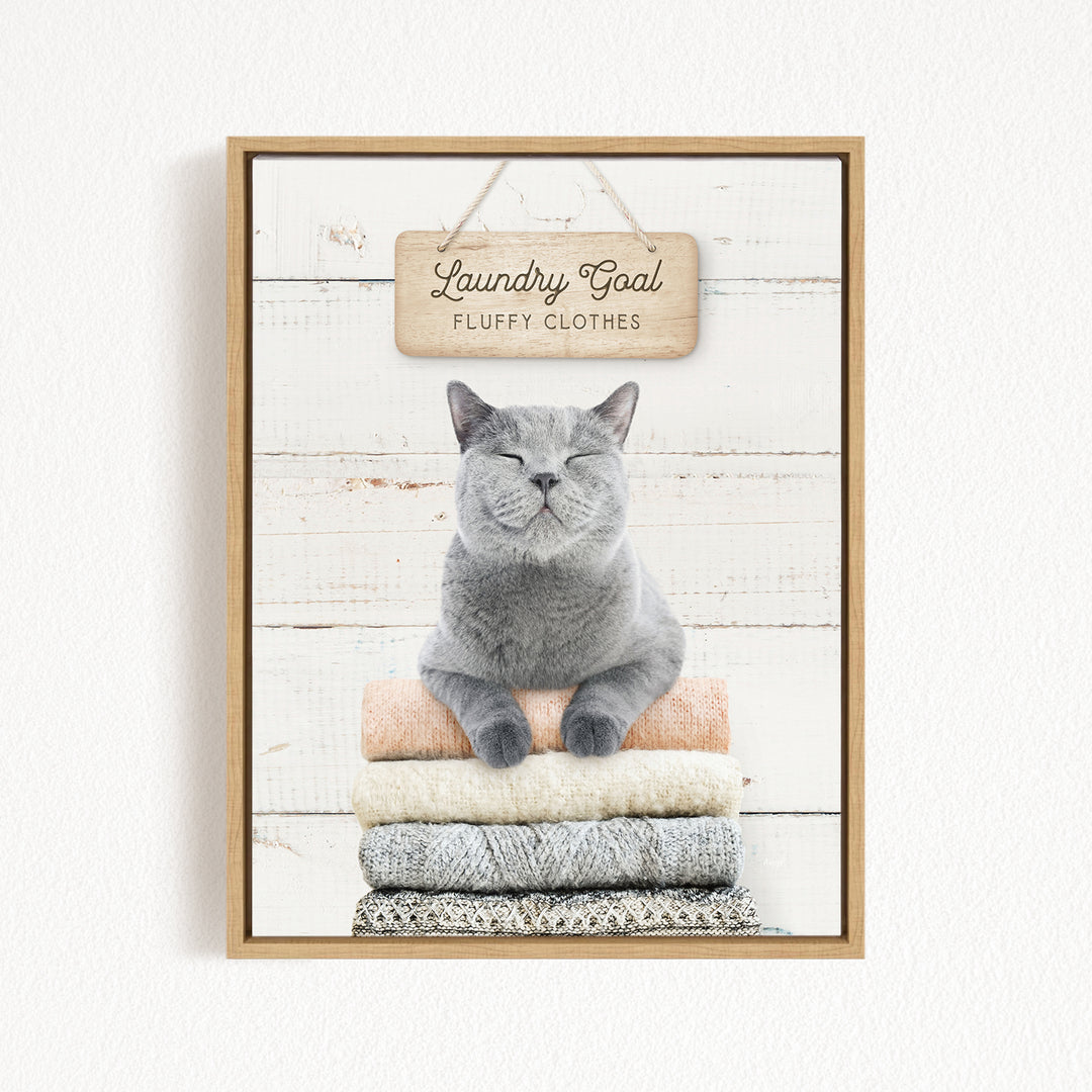 A gray cat sitting on a stack of clothes with a sign above it that says "Laundry Goal Fluffy Clothes".