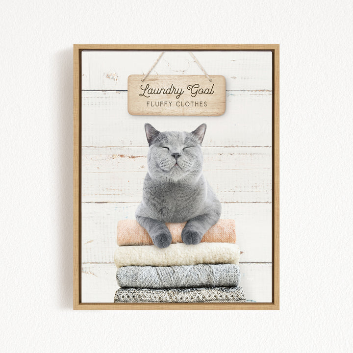 A gray cat sitting on a stack of clothes with a sign above it that says "Laundry Goal Fluffy Clothes".