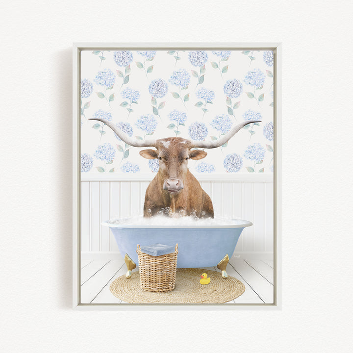 A brown steer with large horns is sitting in a blue bathtub filled with bubbles, surrounded by a wicker basket and rubber duckies.