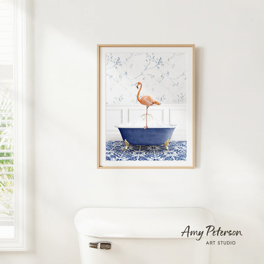 A framed painting of a pink flamingo standing on a blue bathtub is hanging on a white wall in a bathroom.