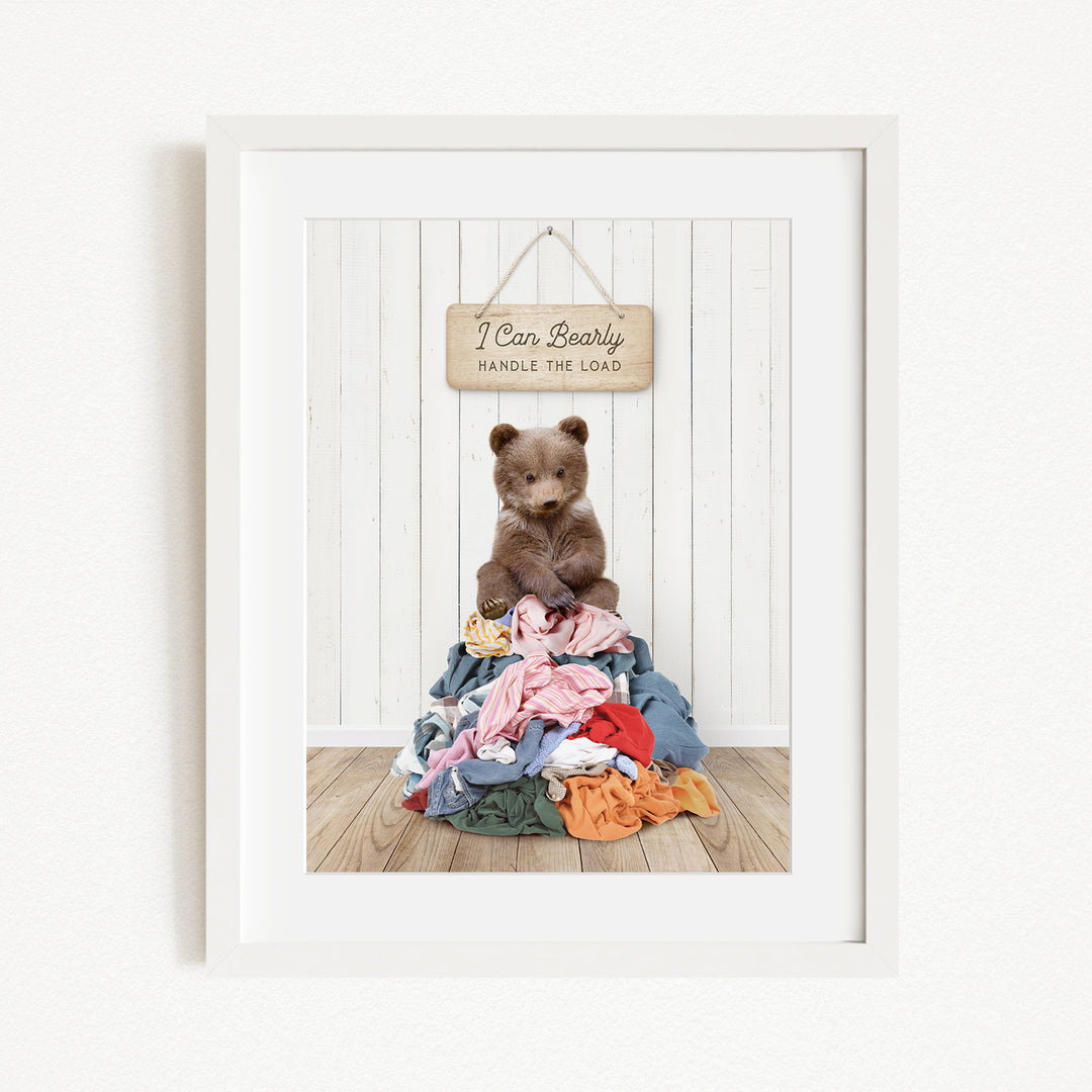 A framed image of a brown bear sitting atop a pile of clothes, with a wooden sign above that reads "I Can Bearly Handle the Load".
