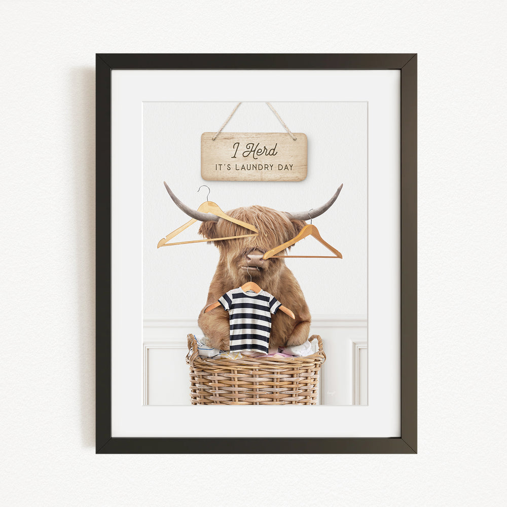 A framed image depicting a Highland cow sitting in a wicker basket, surrounded by clothes and a sign that reads "I herd it's laundry day".