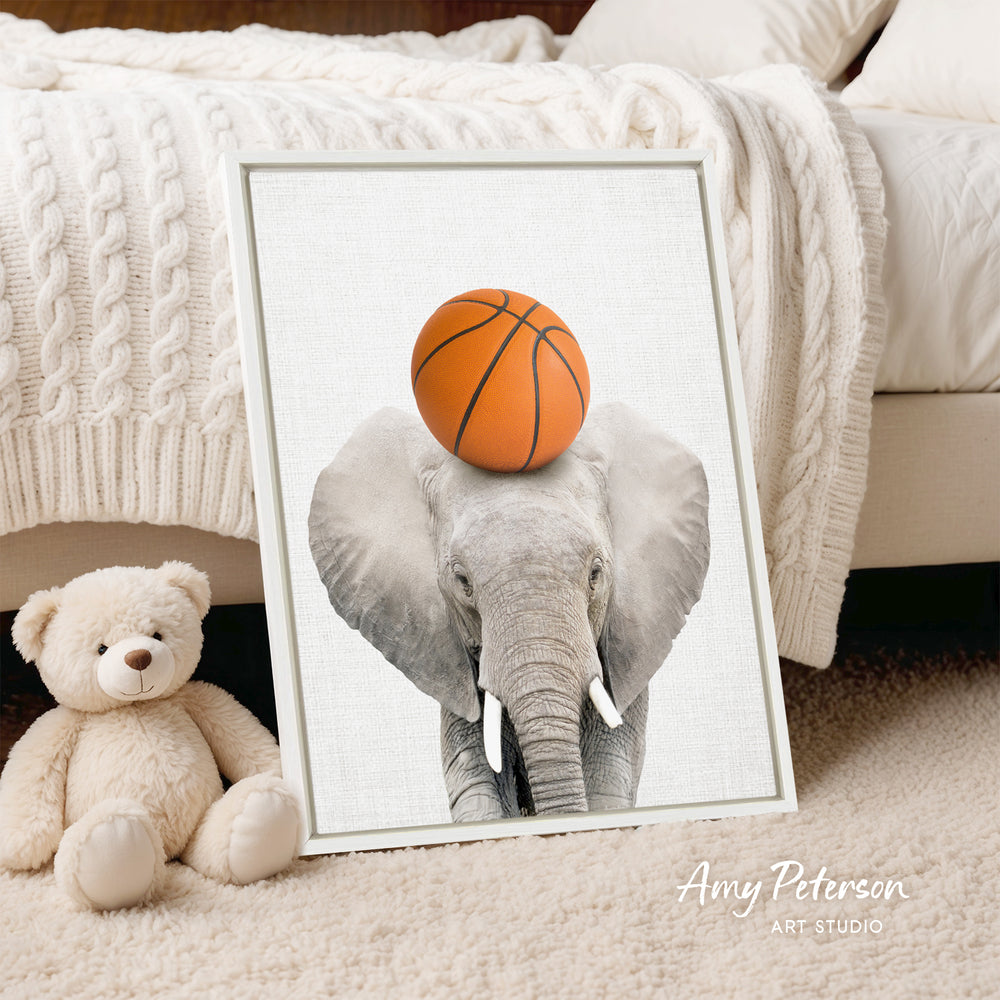 A framed image of an elephant holding a basketball on its head, placed on a bed next to a teddy bear.