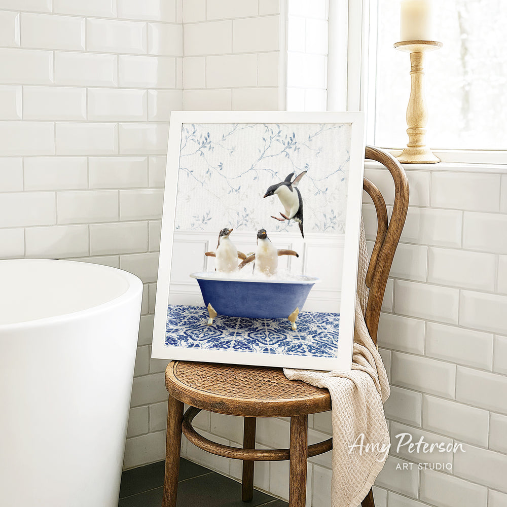 A framed image of penguins sitting in a blue bathtub, with a towel draped over a chair in a bathroom setting.