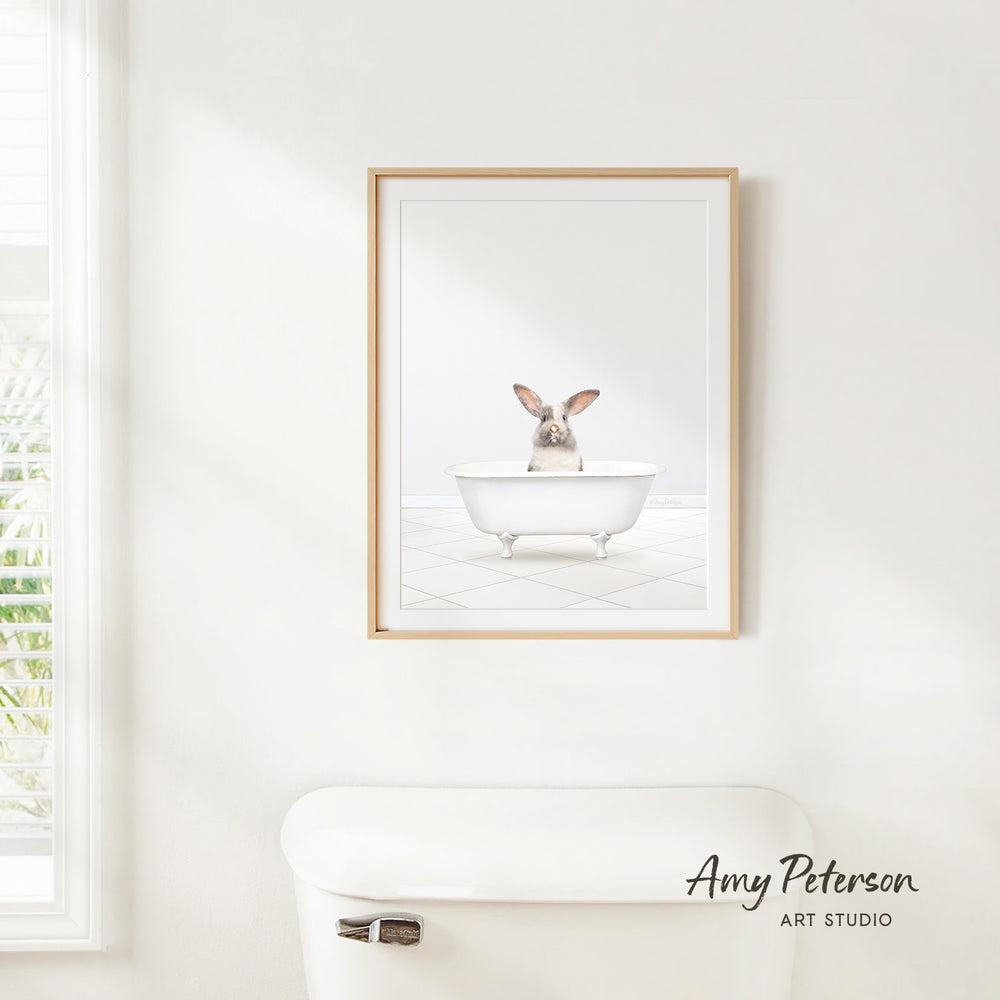 A framed image of a rabbit sitting in a bathtub is hanging on a white wall above a white toilet.