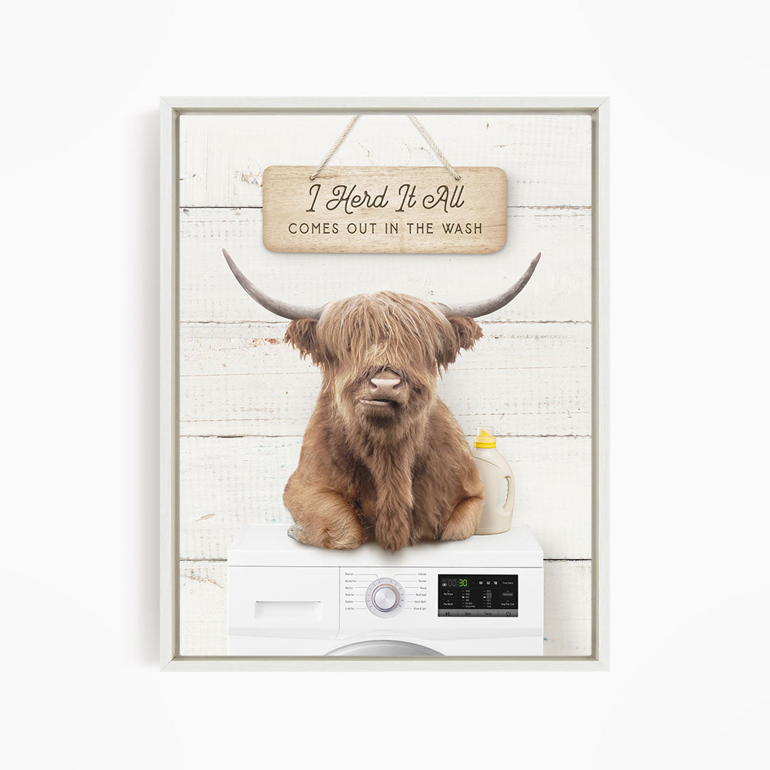 A brown cow with long horns is sitting on top of a white washing machine, with a wooden sign above it that says "I Herd It All Comes out in the Wash".