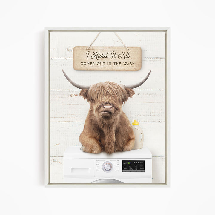 A brown cow with long horns is sitting on top of a white washing machine, with a wooden sign above it that says "I Herd It All Comes out in the Wash".