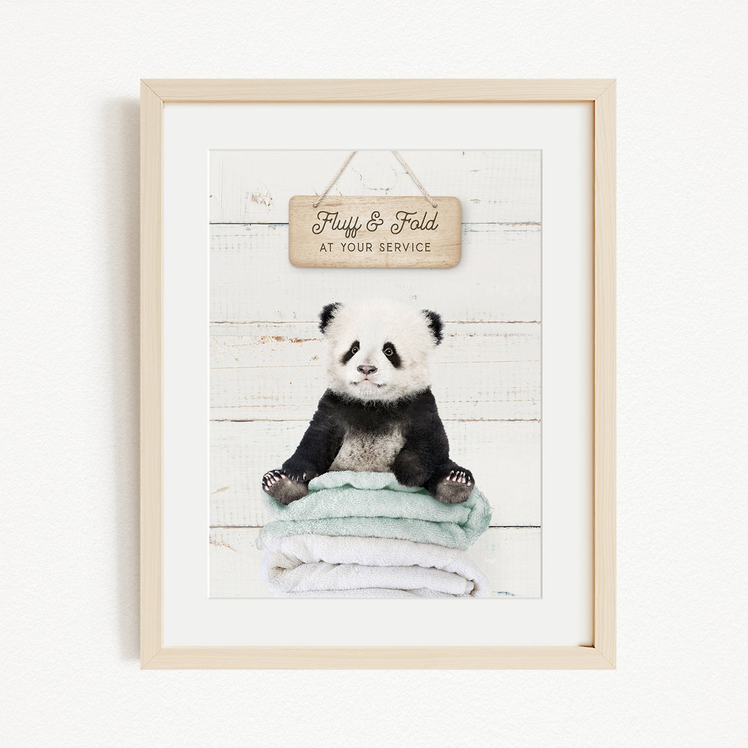 A framed image of a panda bear sitting on a stack of towels, with a wooden sign above that says "Fluffy & Fold at your service".