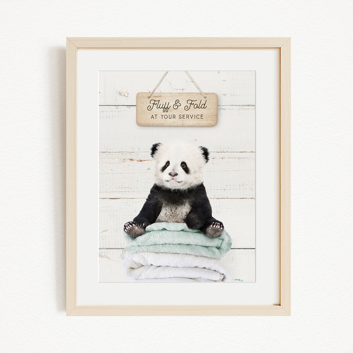 A framed image of a panda bear sitting on a stack of towels, with a wooden sign above that says "Fluffy & Fold at your service".